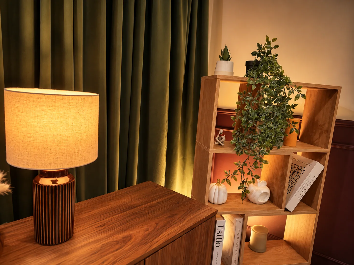 An amber lamp beside wooden shelving with trailing plants and ceramics